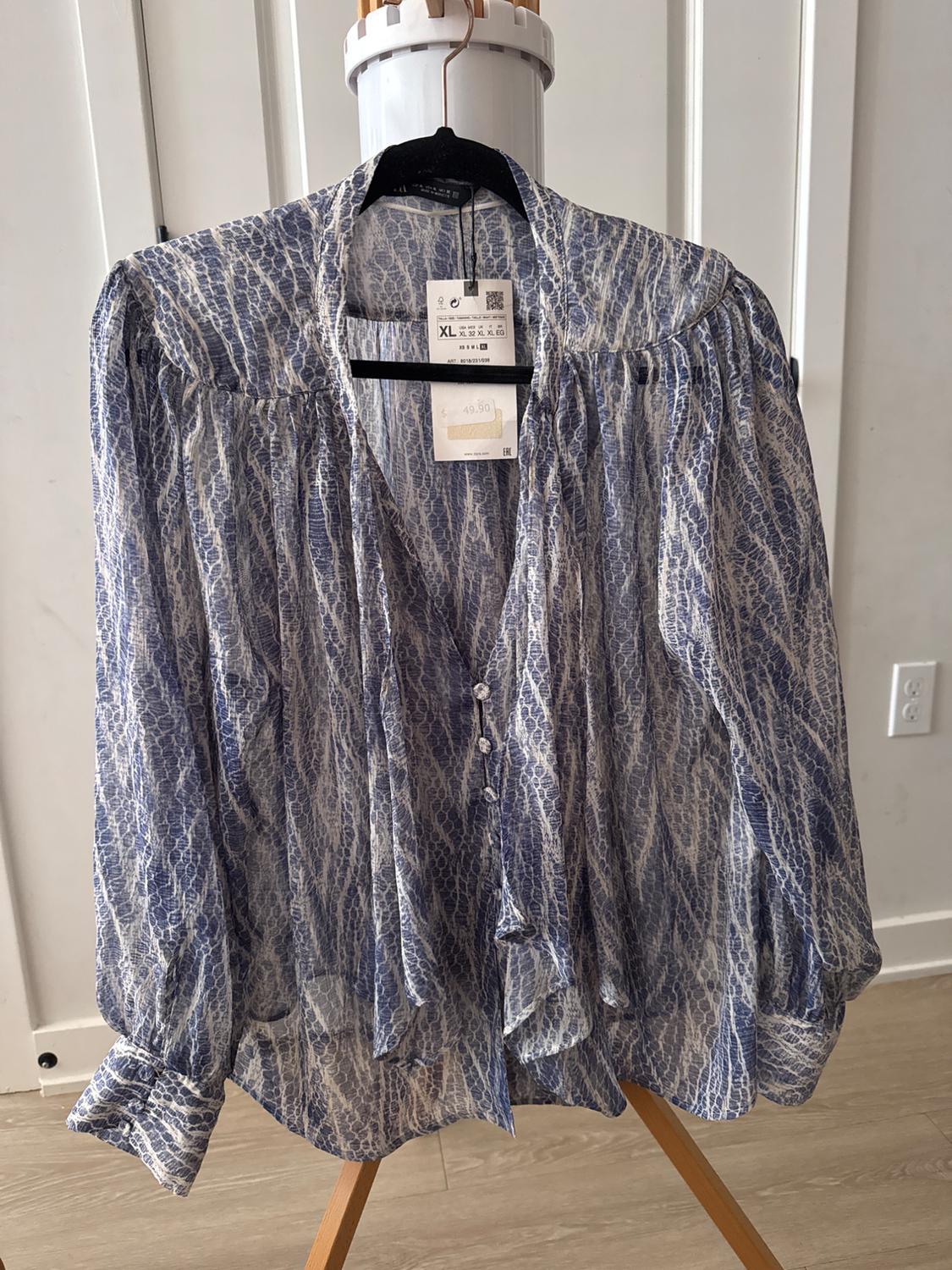 Blue and white patterned blouse with buttons, hanging on a mannequin. Visible label with size XL and price of 49.90. White wall background.