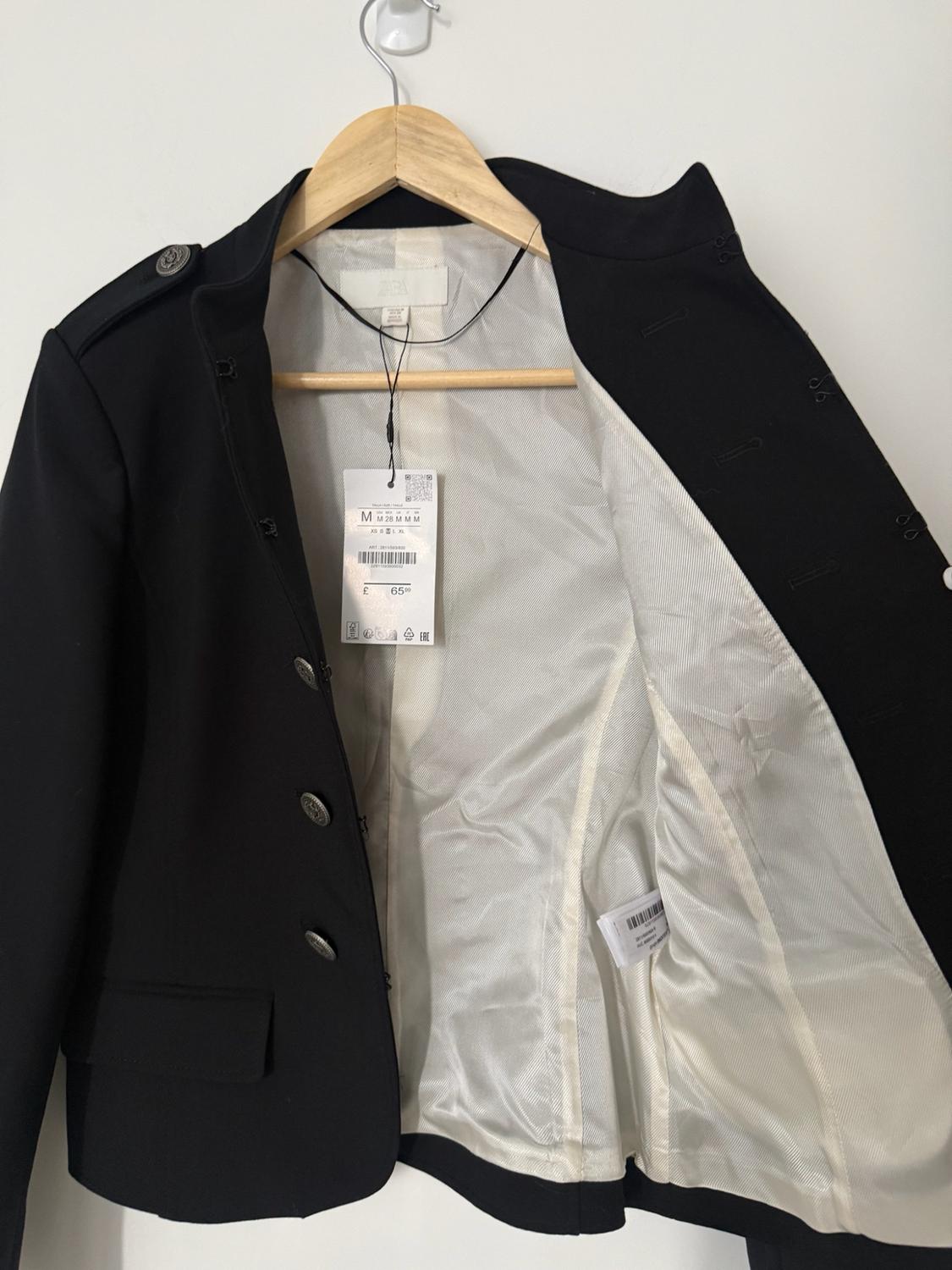 Open black jacket with silver buttons and white lining on a wooden hanger. Visible text: ZARA M M 28 M M M £ 65.99.