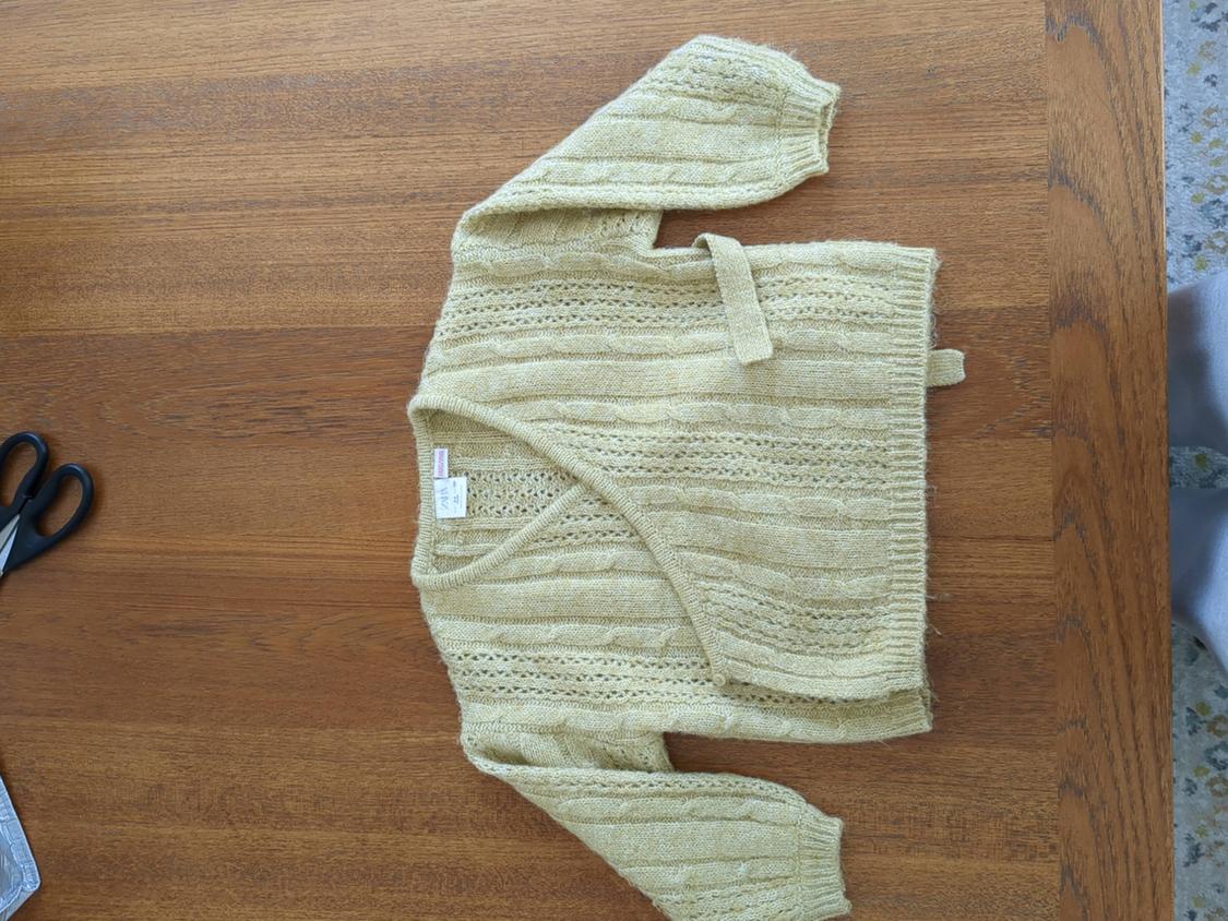 Beige knit cardigan on a wooden table, with black scissors on the left. Visible tag inside the collar.