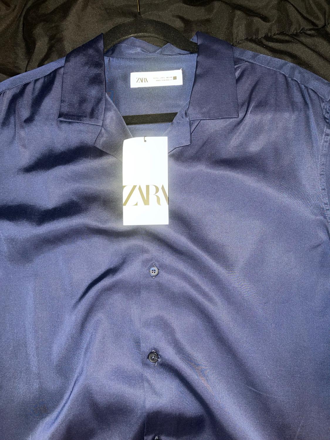Dark blue button-up shirt made of shiny fabric on a black hanger. Shows tags with the text zara. Dark fabric background.