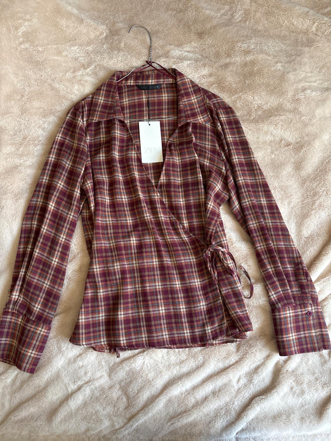Grenadine checked long-sleeved wrap blouse with a tie, on a hanger over a soft blanket. Label with ZARA text.