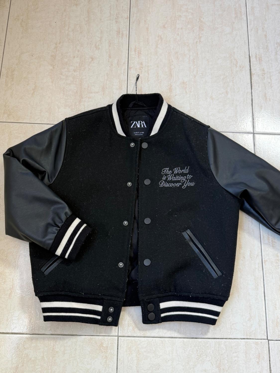 Black varsity jacket with faux leather sleeves, "The World is Waiting to Discover You" text on the chest, on a light-coloured floor.