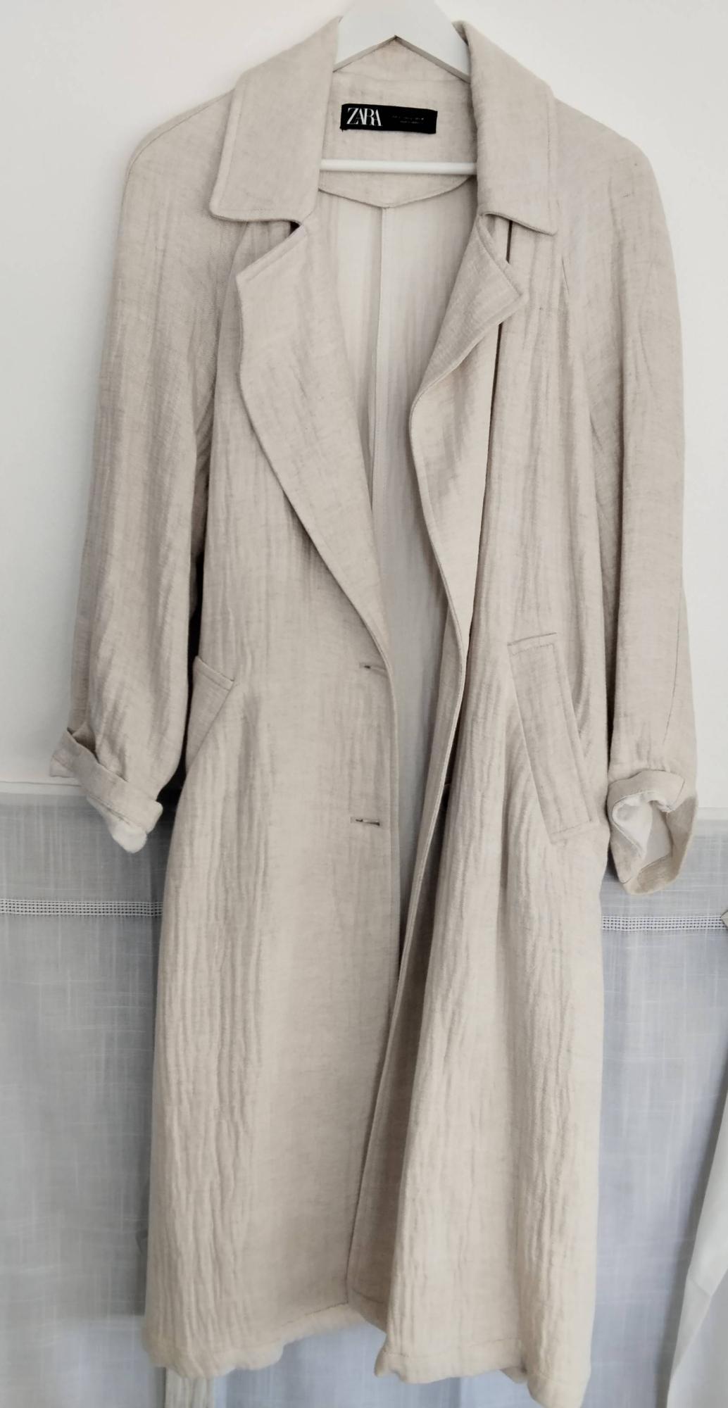 Long beige coat with a lightweight texture hanging on a white hanger against a light background. Its lapels, pockets and ZARA label stand out.