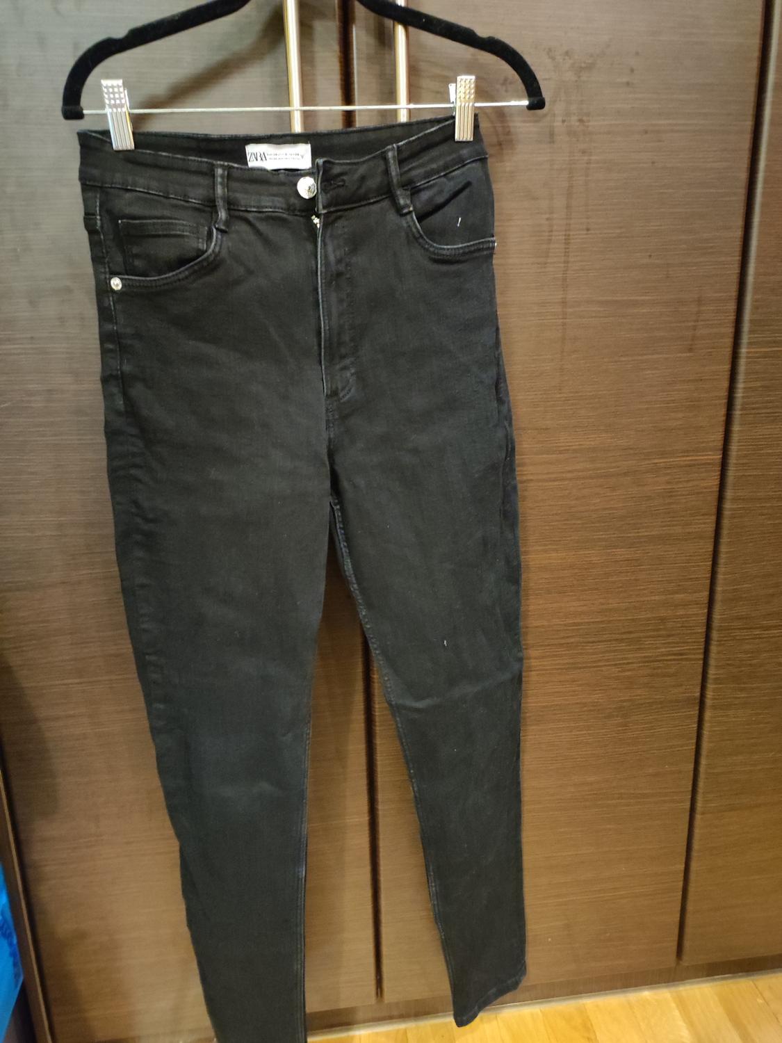 Black trousers hanging on a hanger in front of a wooden wardrobe. Label visible at the waist.