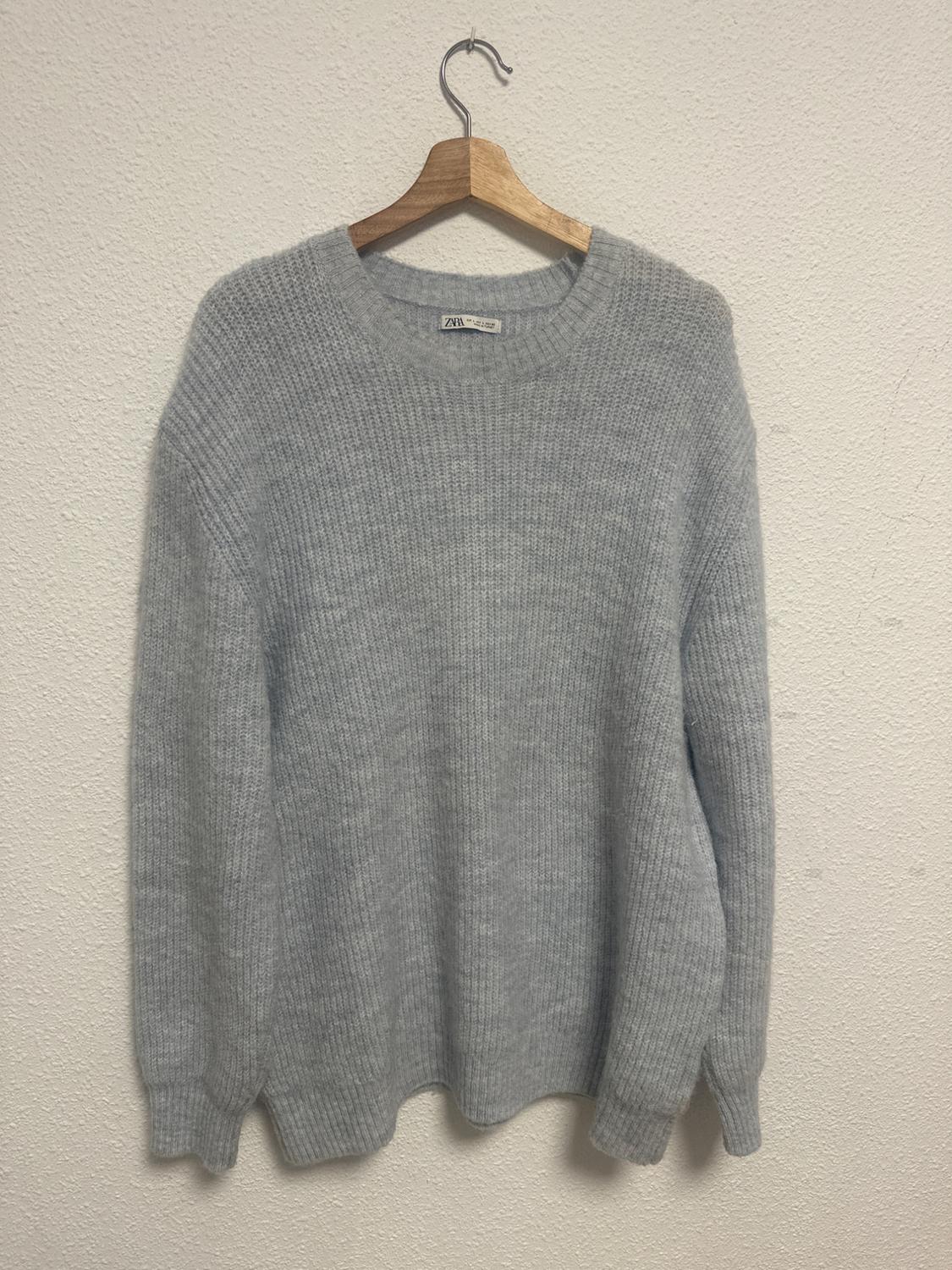 Chunky grey knitted jumper hanging on a wooden hanger against a white wall. Visible brand label.
