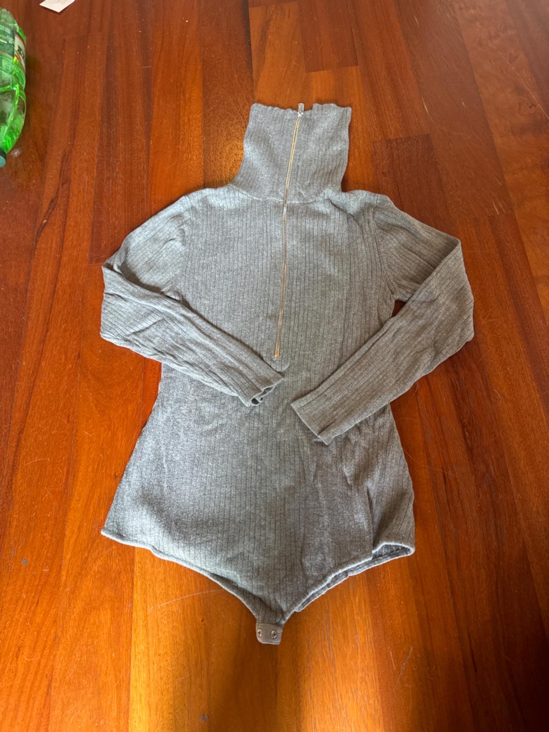Grey long-sleeved bodysuit with front zip, on a wooden floor. Green bottle visible in the top left corner.