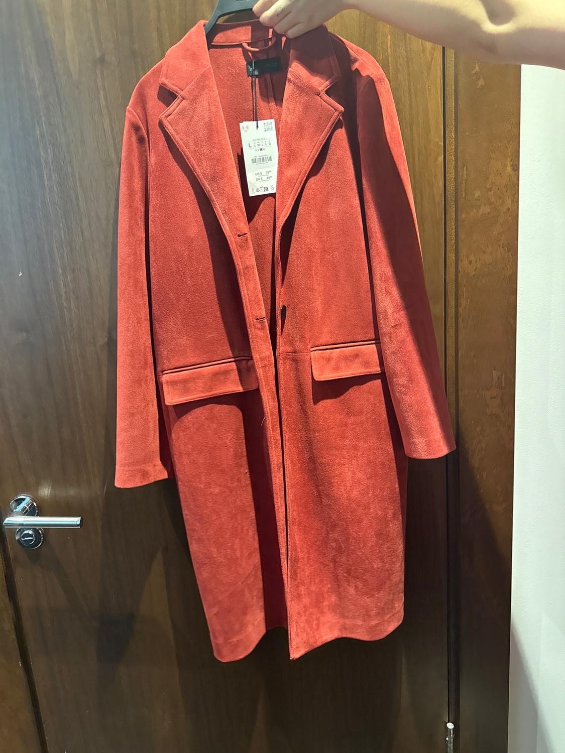 A red coat is hanging on a hanger, held by a hand. The product tag is visible. The background is a wooden door.