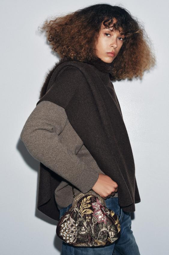 Hnědá kabelka se sadou s květinovou výšivkou a korálky, kovovým uchem a klipovým zapínáním, hnědý svetr a džíny.