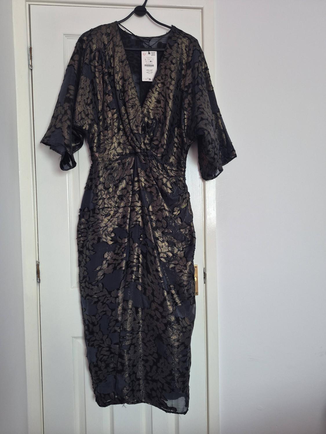 Dark dress with a gold floral print, V-neck, short sleeves and a gathered waist, hanging on a white door.