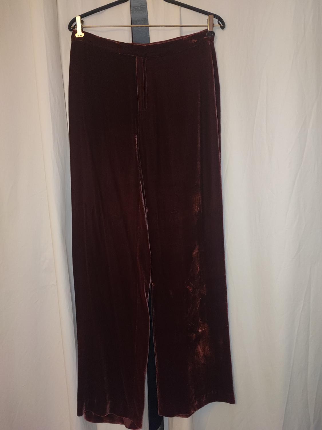 Burgundy velvet trousers hanging on a black hanger against a backdrop of white curtains.