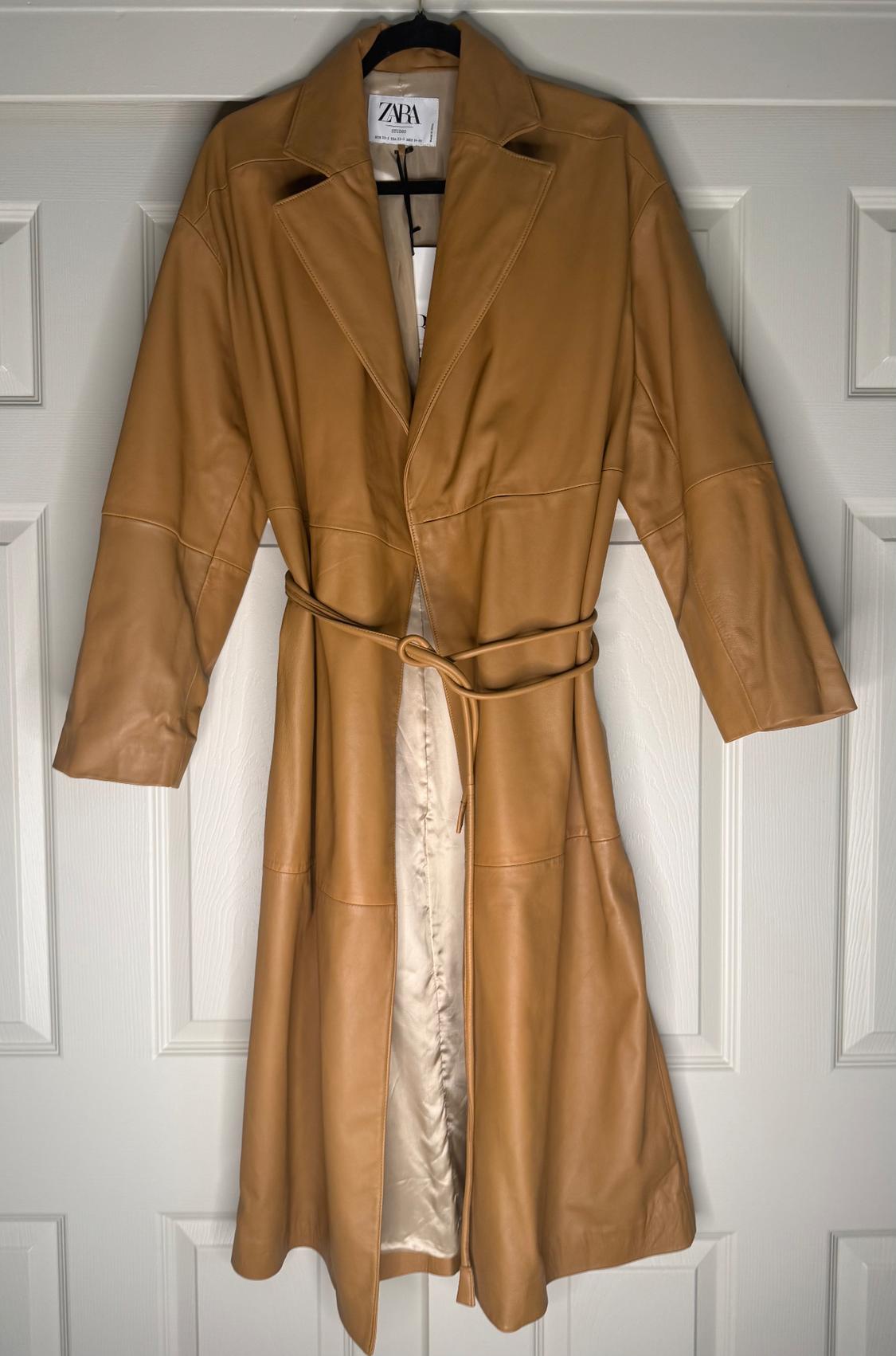 Light brown long coat hanging on a hanger, with a belt of the same material. Visible label with the text "ZARA". White door background.