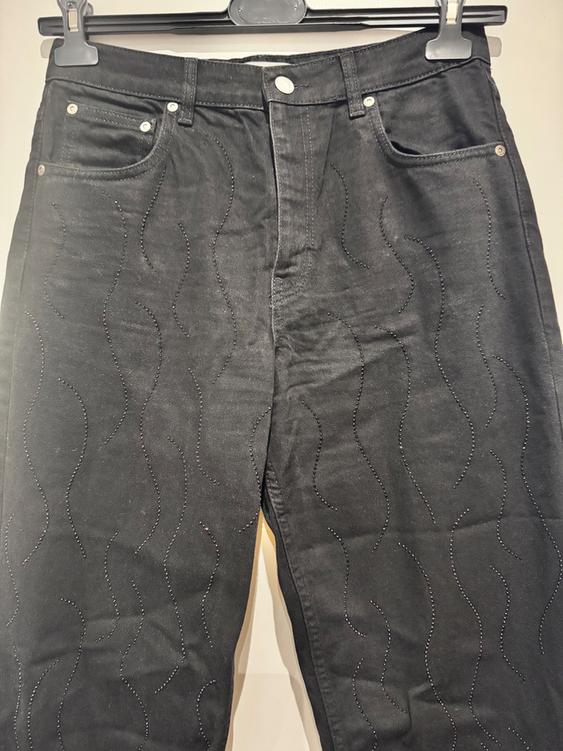 Black trousers with a shiny wavy line design, hanging on a grey hanger against a light background.