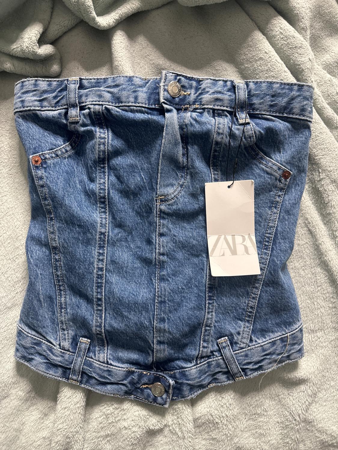 Short blue denim skirt with visible stitching, front pockets, and a white label with the Zara logo, on gray fabric.