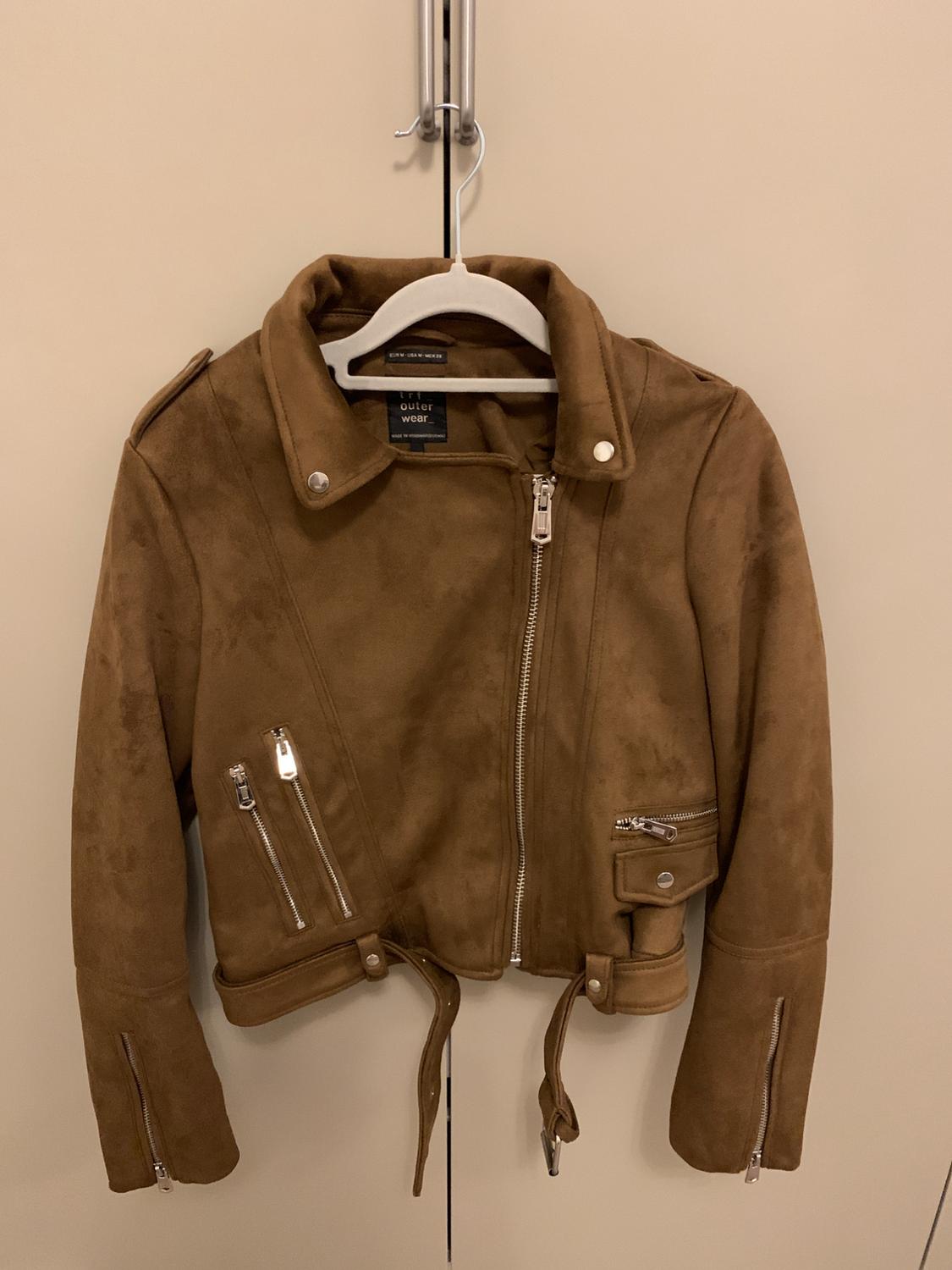 Brown suede jacket with zips and a belt, hanging on a white hanger in front of a beige door. Tag: "outer wear".
