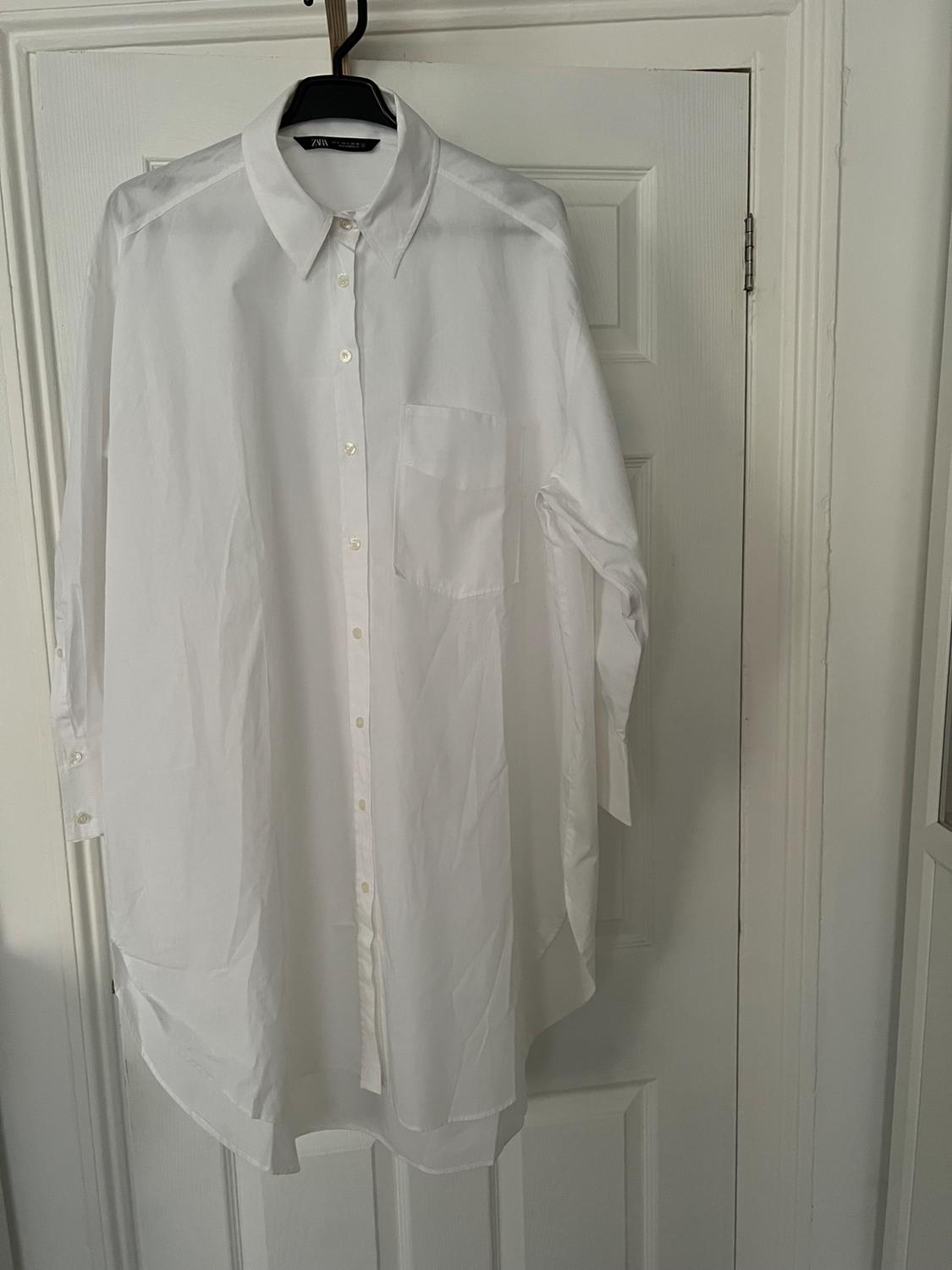 Oversized white long-sleeved shirt with a pocket, hanging on a hanger on a white door. The label on the collar reads ZARA.