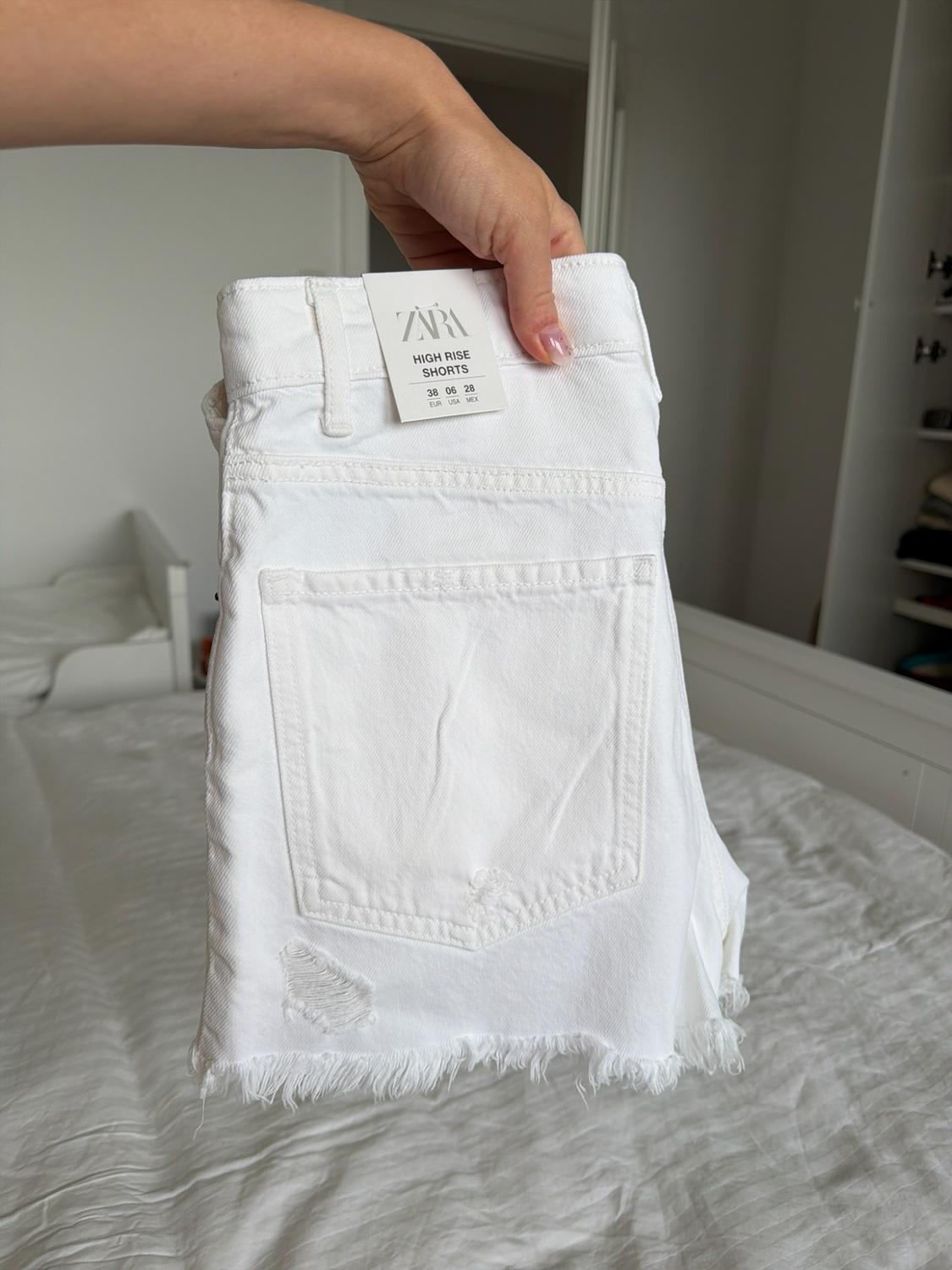 Χέρι κρατάει λευκά σορτς με ξέφτια. Ετικέτα: ZARA HIGH RISE SHORTS 38 06 28 EUR USA MEX. Φόντο κρεβατοκάμαρας με κρεβάτι, casual στυλ.