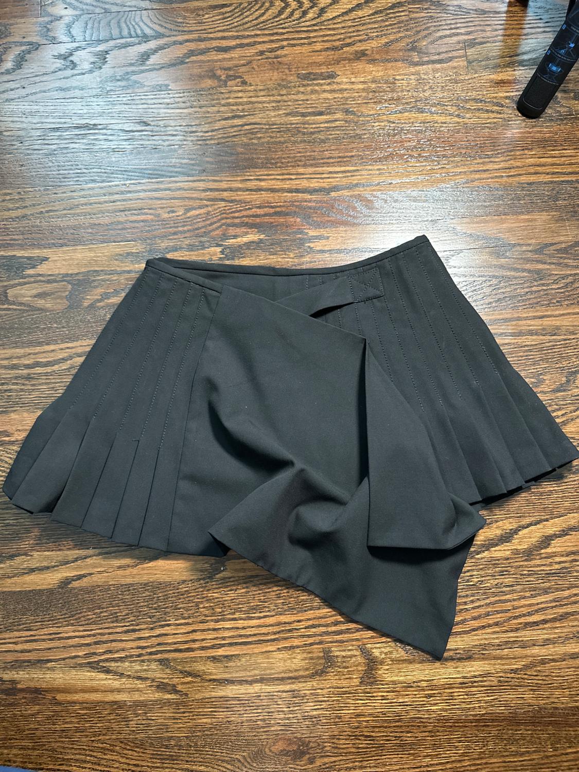 Black pleated skirt on a brown wooden floor. The fabric is slightly wrinkled in the center.