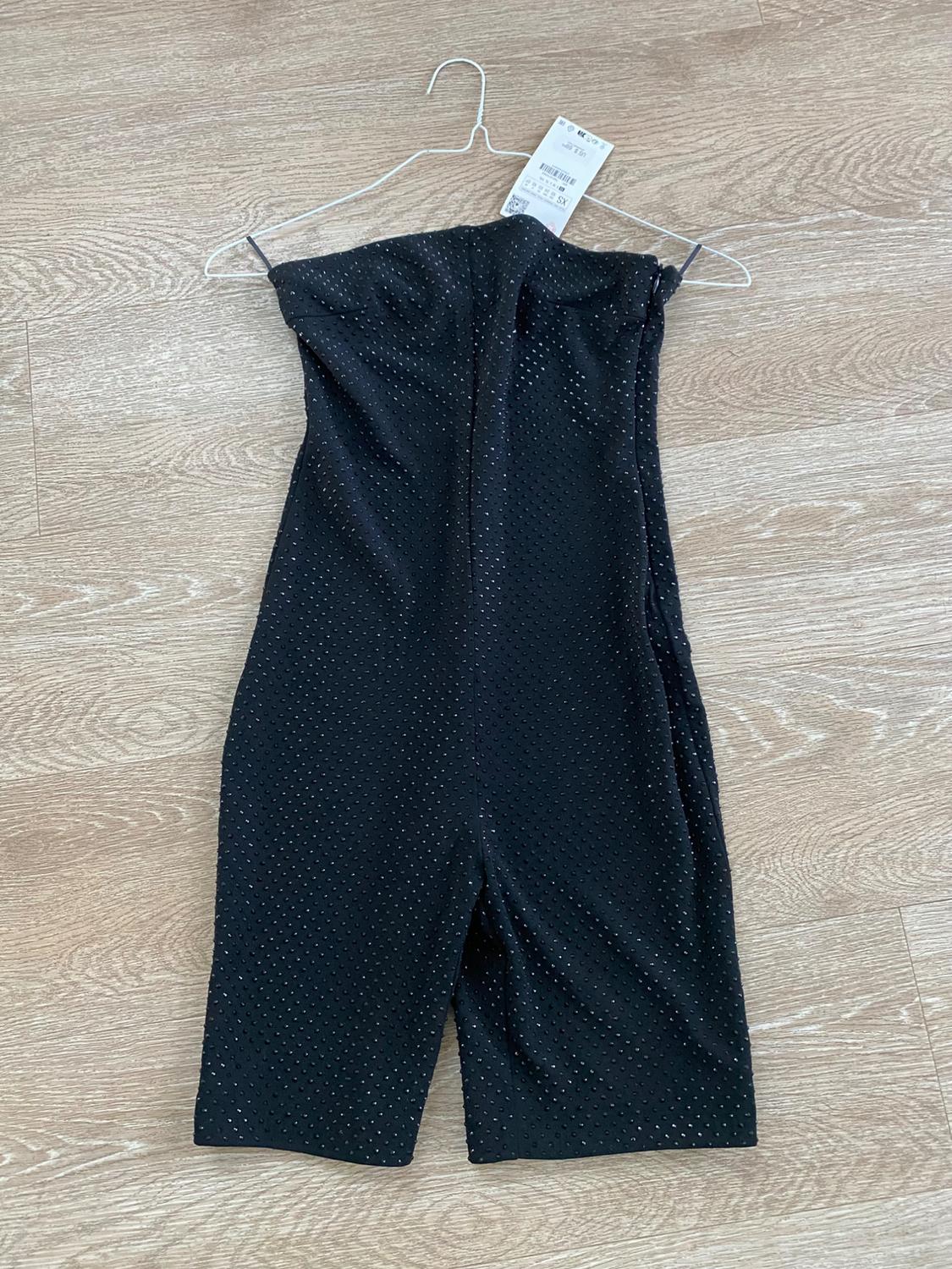 Black jumpsuit with small shiny dots hanging on a hanger over a light wood floor. Tag visible on the hanger.