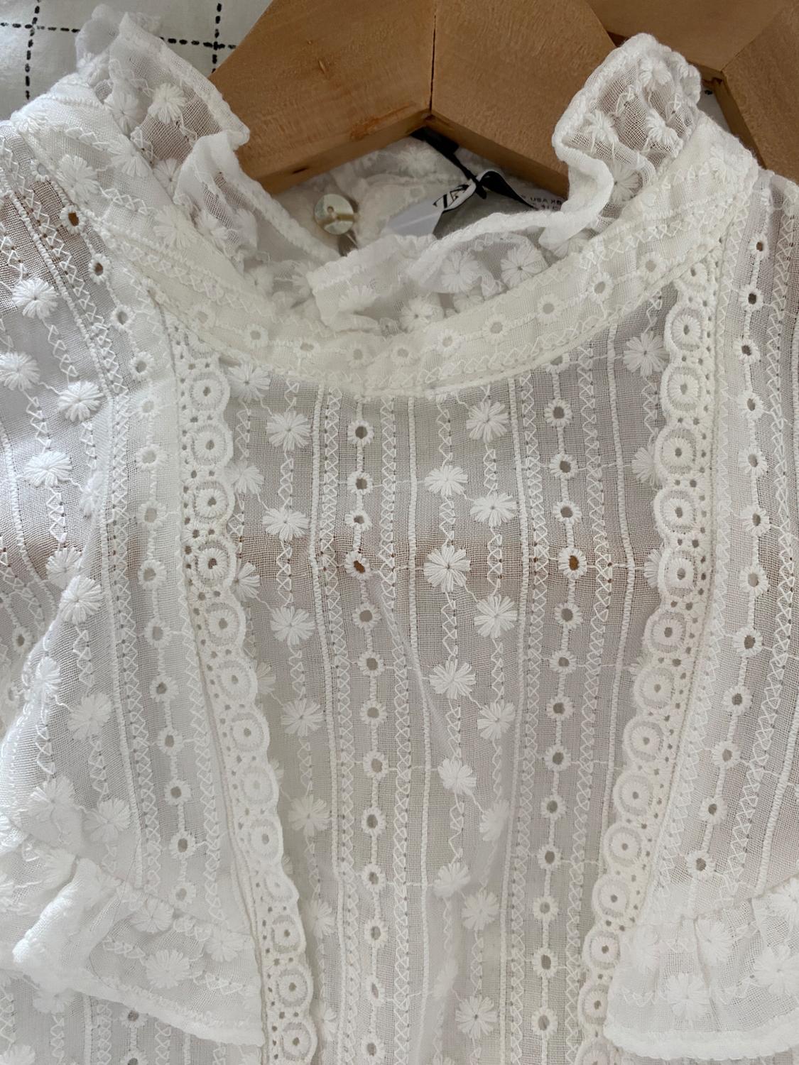 White lace blouse with floral details and embroidery, hung on a wooden hanger against a white checked fabric background.