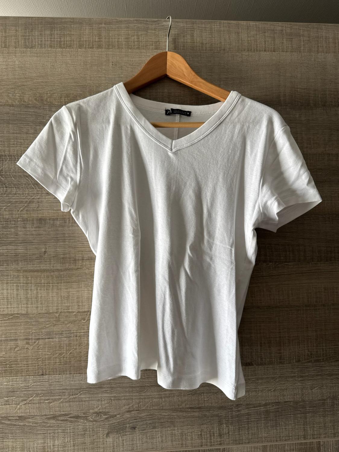 White short-sleeved V-neck T-shirt hanging on a wooden hanger against a wooden wall background. Casual and relaxed style.