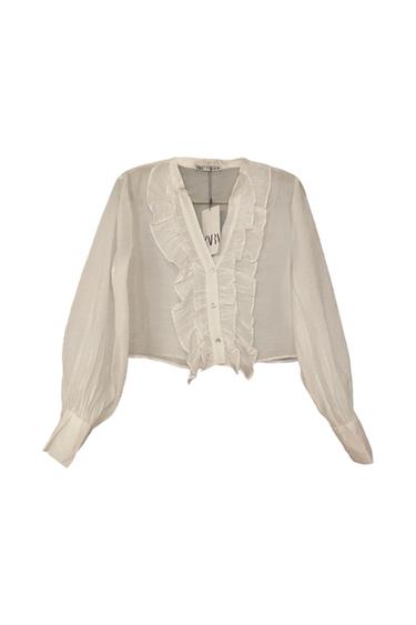 White blouse with ruffles on the front and puffed long sleeves. Visible tag with text "ZARA". White background.