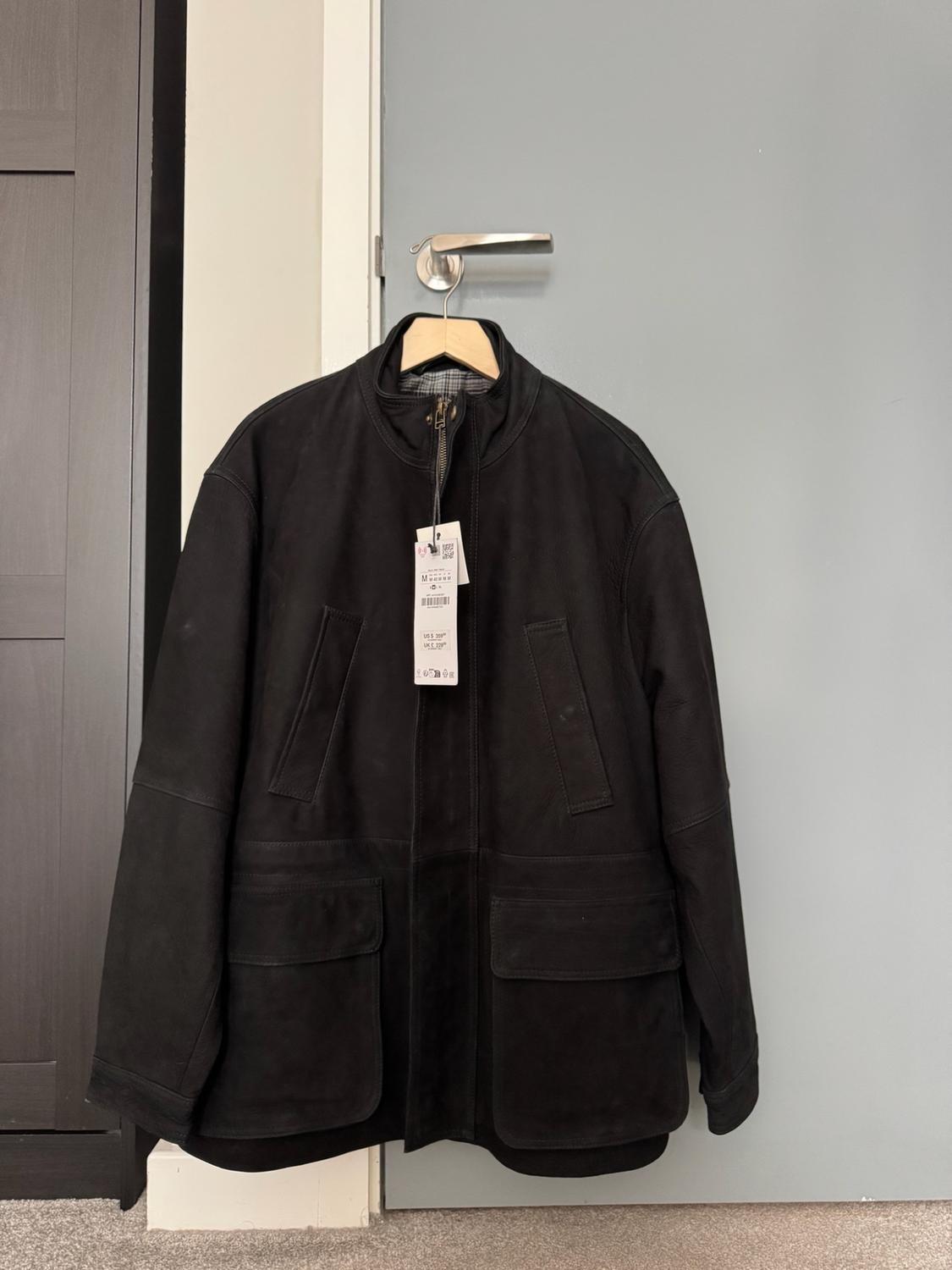 Black jacket with a zip and front pockets, hanging on a wooden hanger from the handle of a grey door indoors.