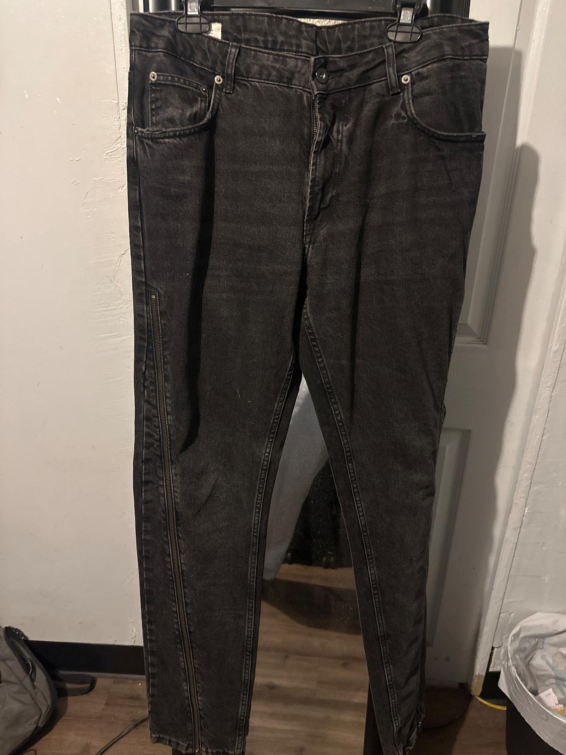 Black jeans hanging on a hanger, with visible seams and a white wall and wooden floor background.