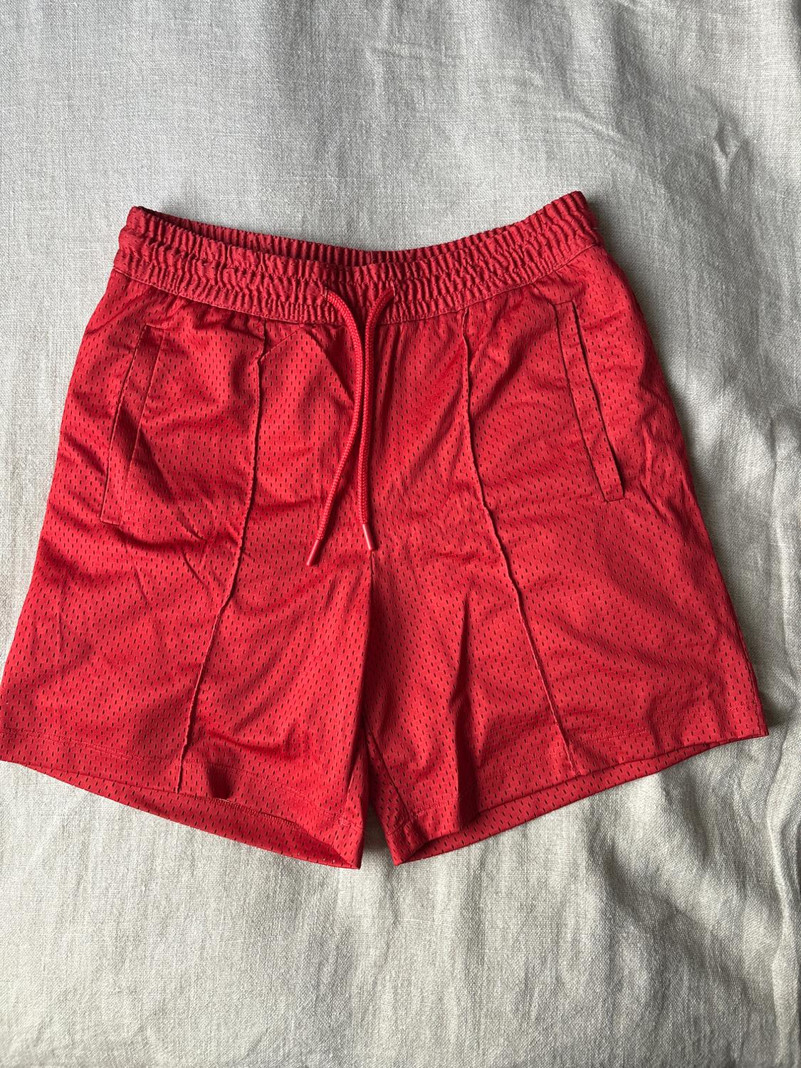 Red perforated fabric shorts with an elasticated drawstring waist, on a grey fabric surface.