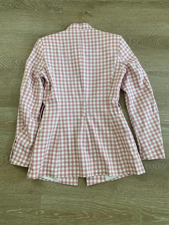 Pink and white gingham jacket hanging over a wooden floor. Back view, with a fitted cut and an opening at the bottom.