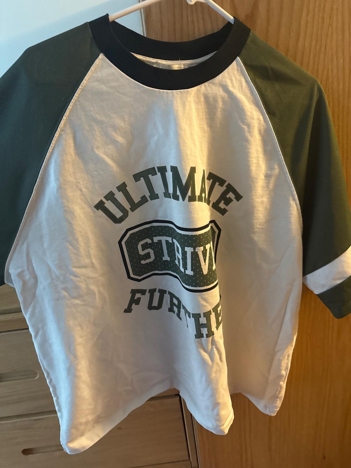 White t-shirt with green sleeves hanging on a hanger. Text on the front: "ULTIMATE STRIVE FURTHER". Light wood background.