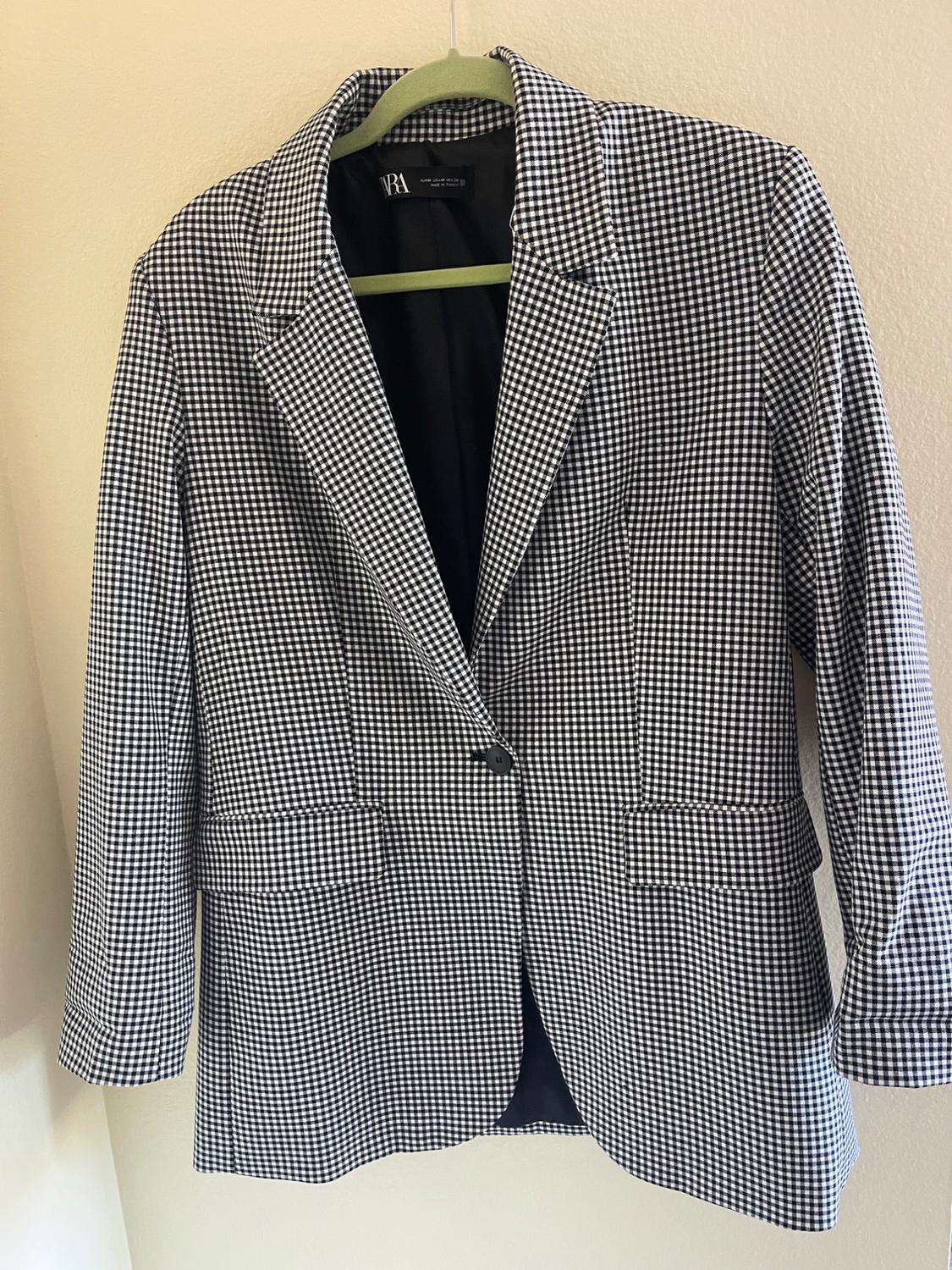 Black and white checked jacket hanging on a green hanger. Visible label with the text "ZARA". Beige wall background.