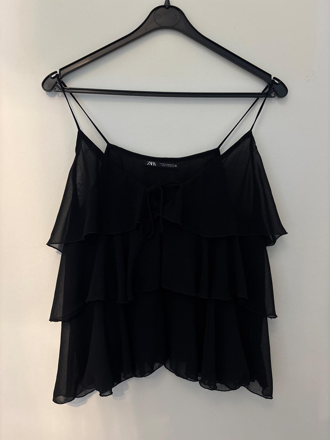 Black strappy top with layered ruffles, hanging on a hanger against a white wall. Text on label: ZARA.