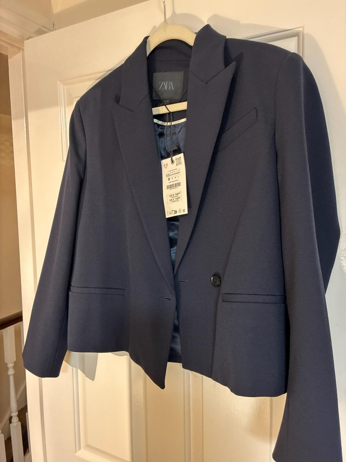 Navy blue blazer hanging on a white door. Visible labels with the text ZARA, XS, US $ 109, UK £ 109.