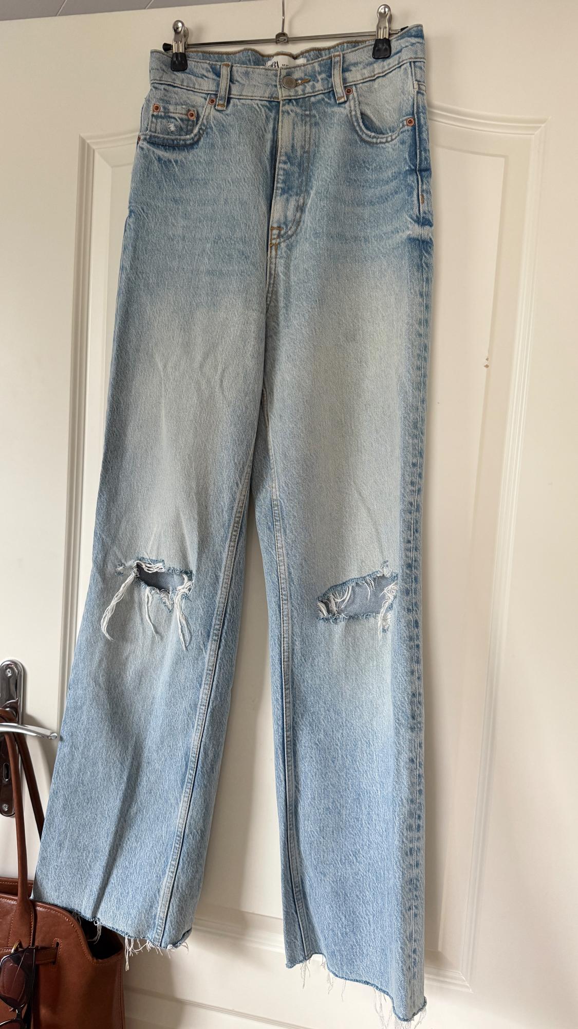 Distressed blue jeans with rips on the knees, hanging on a white door. Back pocket visible.