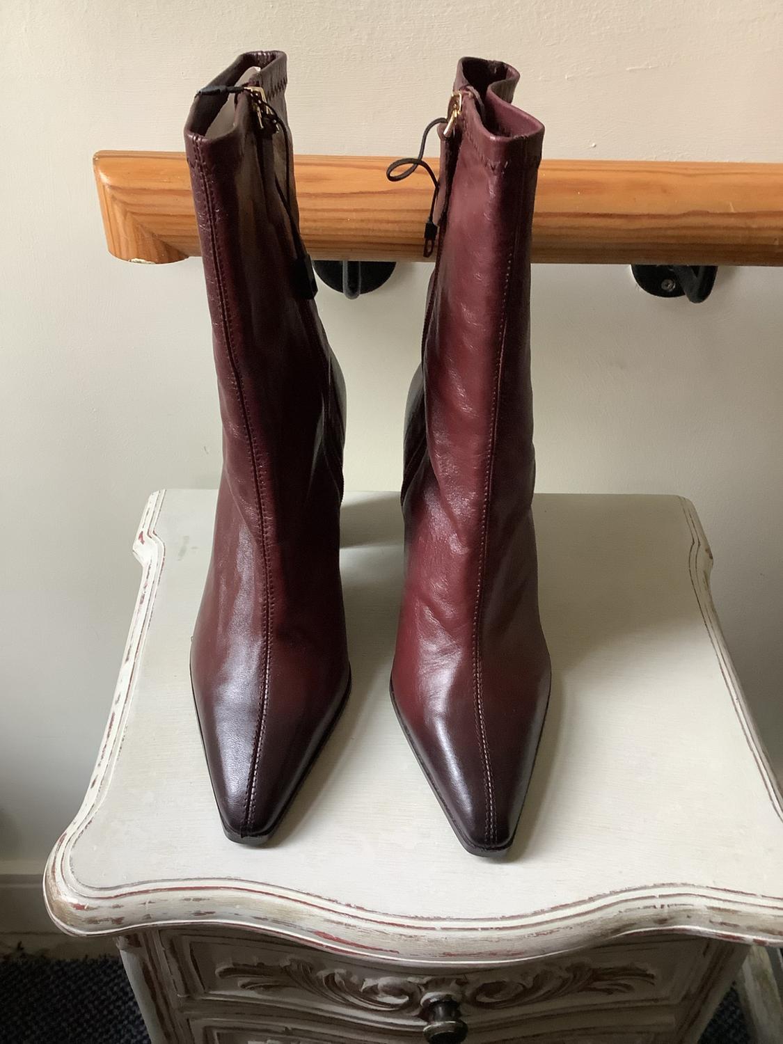 A pair of burgundy pointed ankle boots on a distressed white side table. In the background, a light-coloured wall and a wooden handrail.