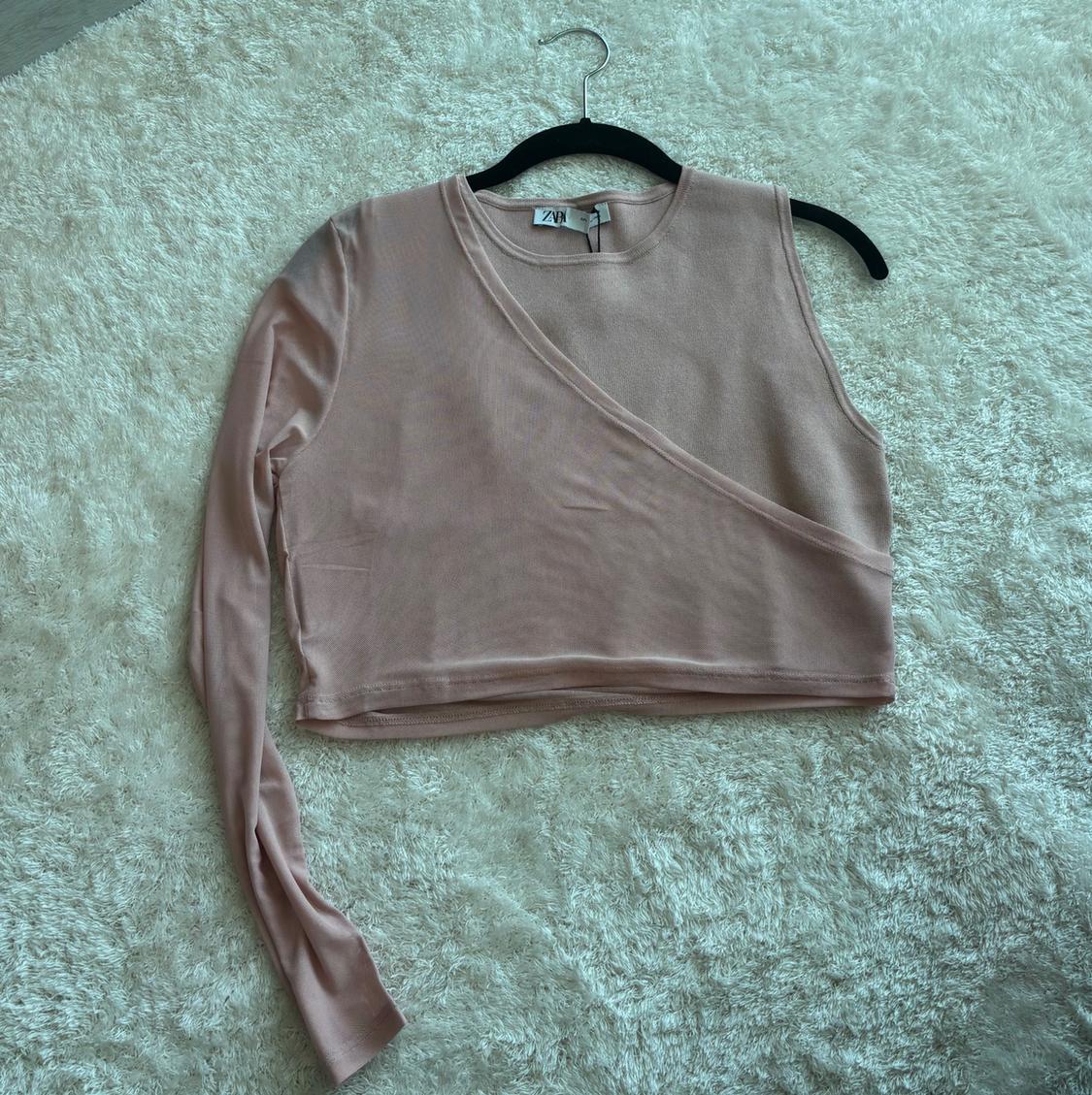 Light pink asymmetric top with a long sleeve, hanging on a black hanger over a beige carpet.