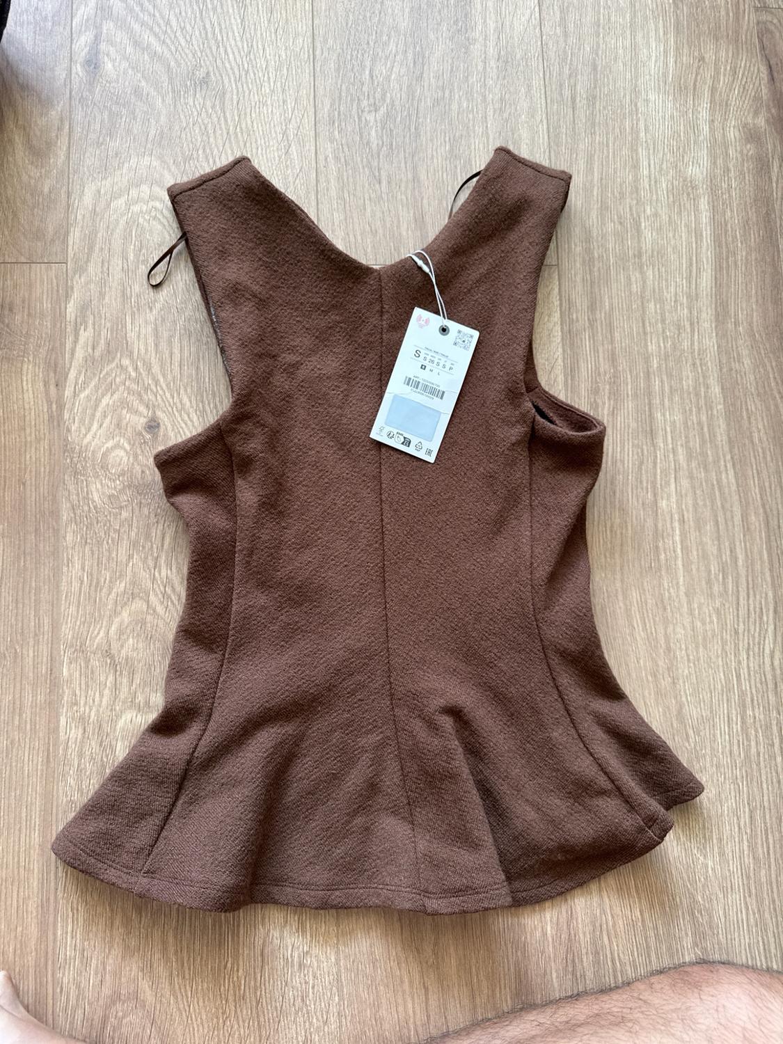 Rear view of a sleeveless textured brown top, with a peplum hem and size S label, on a wooden floor. A leg is visible.