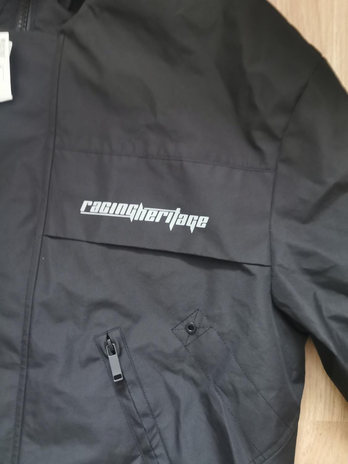 Black jacket with zip fastening and button on the pocket. "racingheritage" inscription on the chest. Light wooden background.