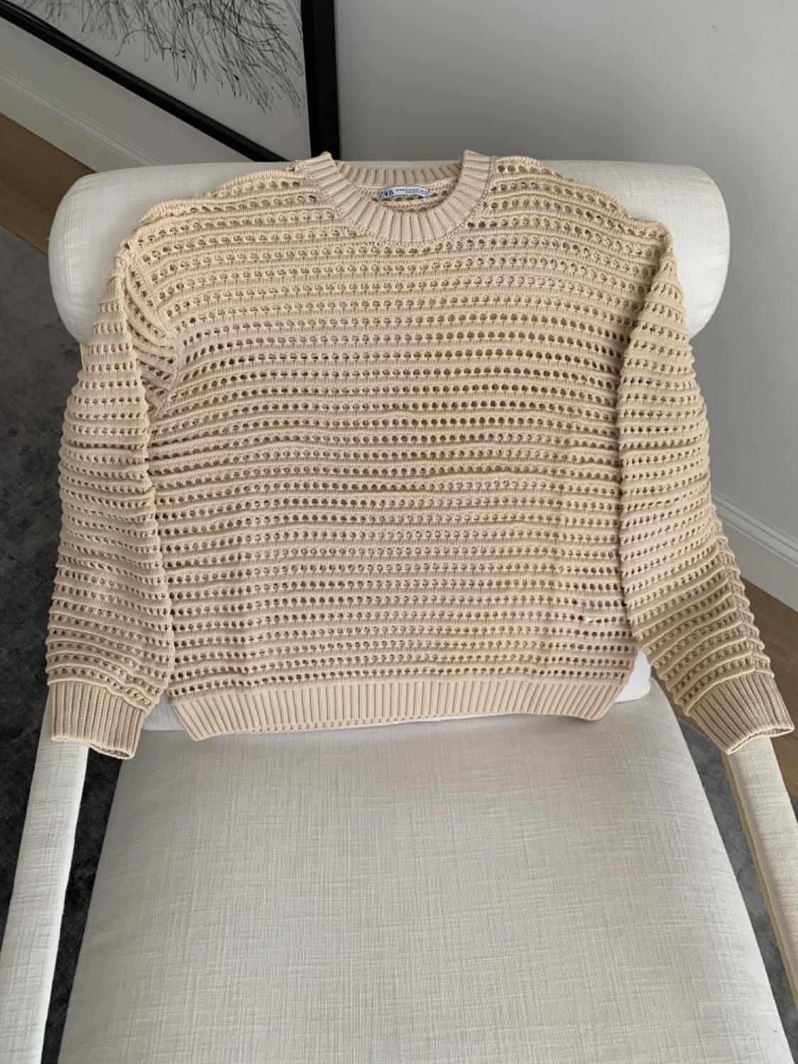 Beige open knit jumper draped over a white chair in a minimalist setting with a picture on the wall in the background.