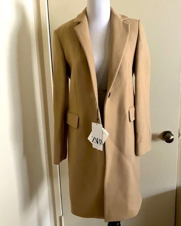 Camel coat on a white mannequin, with a visible label that says "ZARA", in an indoor setting with a door in the background.