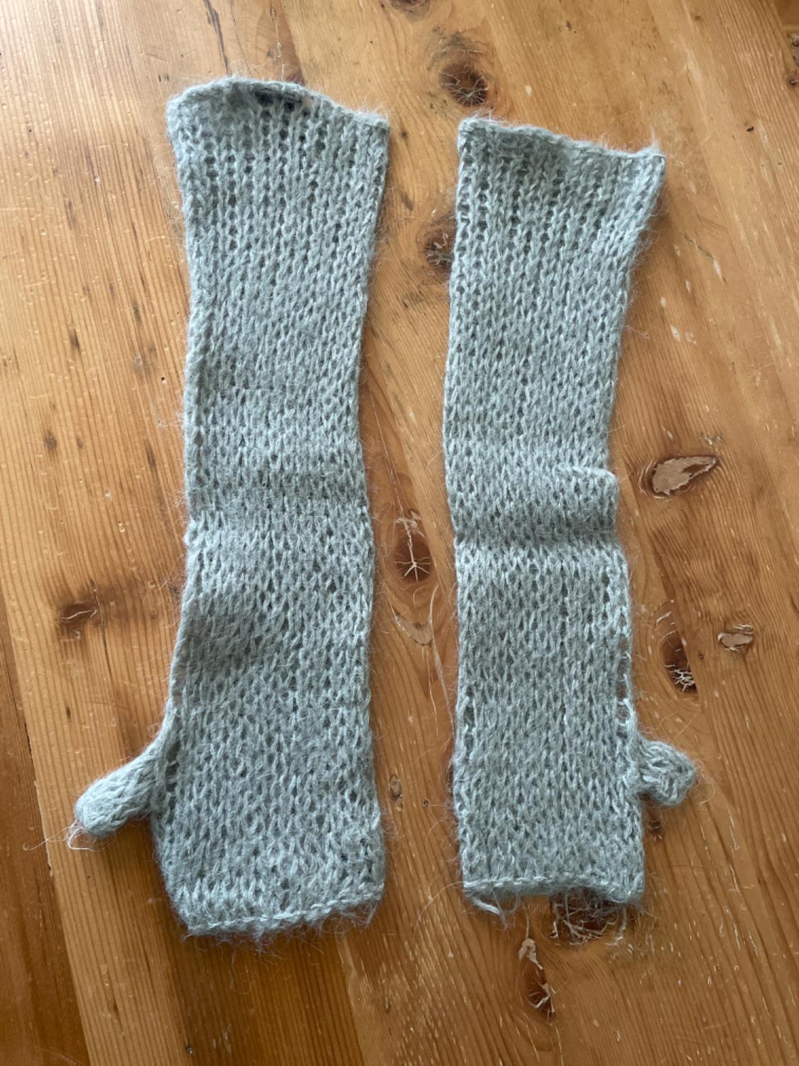 Two light grey fingerless knitted gloves on a wooden table. The gloves have a simple design and a soft texture.