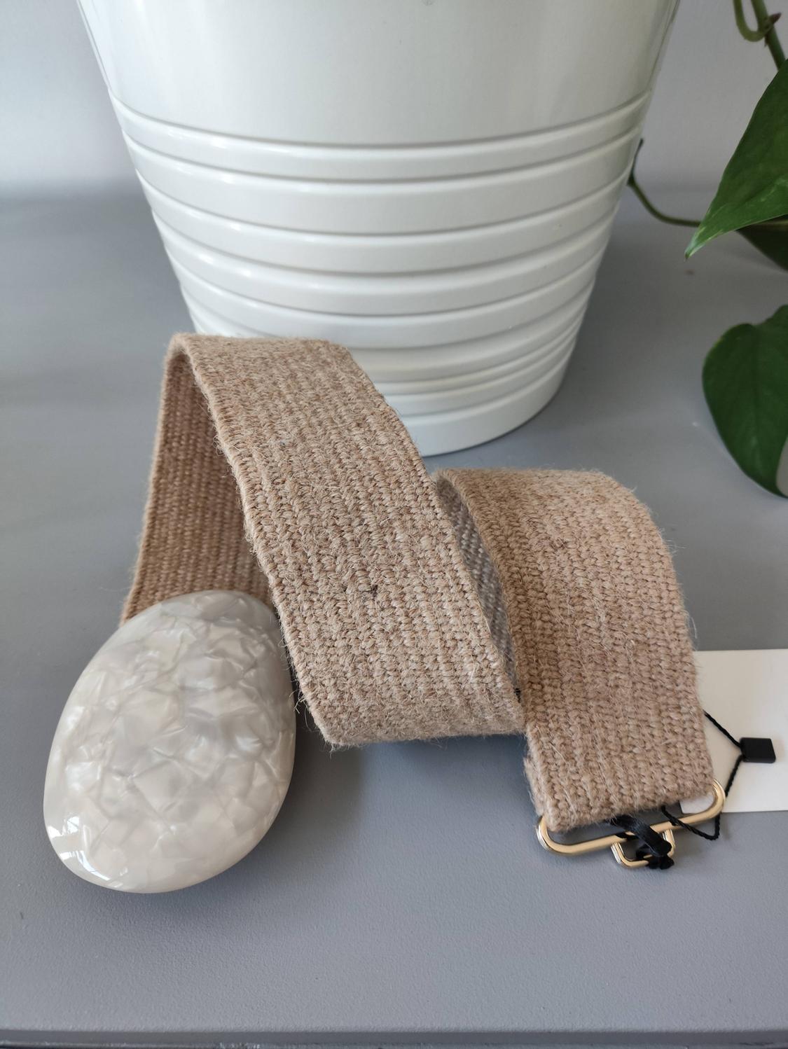 Beige woven belt with a large white marble-effect oval buckle, on a grey surface next to a white plant pot and green leaves.