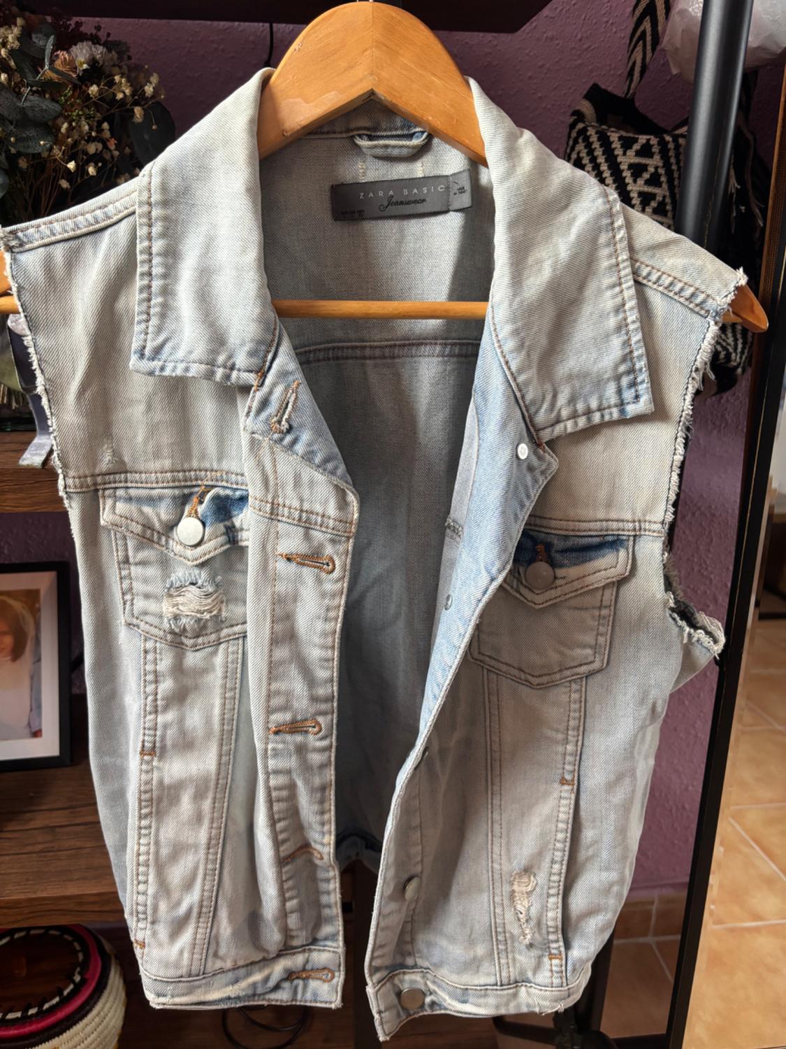Light blue distressed denim waistcoat hanging on a wooden hanger, with a bookshelf and decor in the background.