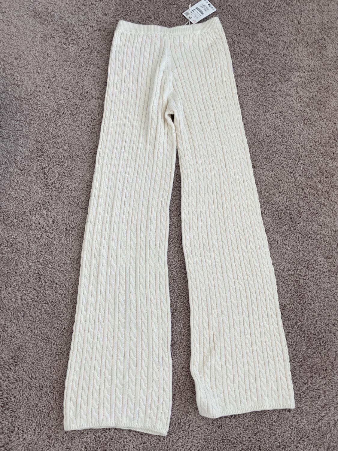 Cream-colored cable knit pants on a gray rug, with a visible label on the waistband.