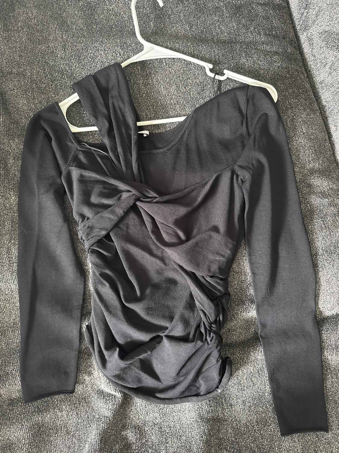 Long-sleeved black blouse with draped design and asymmetrical neckline, hanging on a white hanger against a gray background.