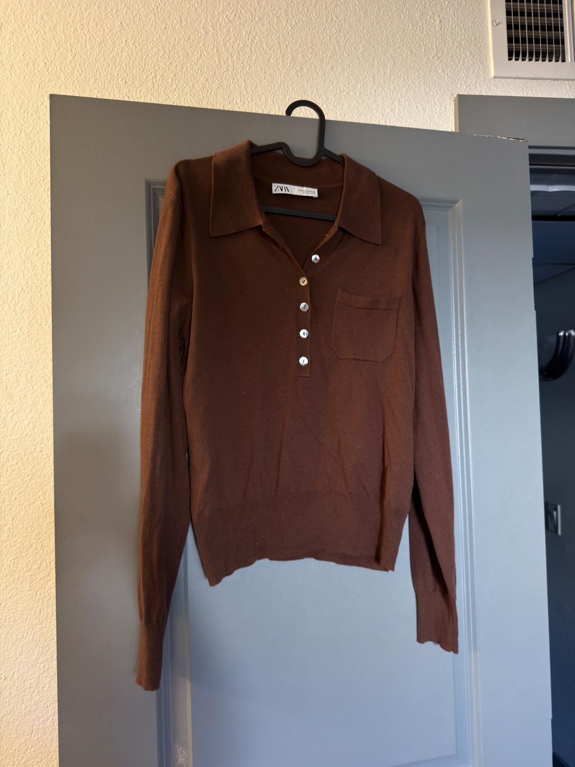 Brown long-sleeved knitted polo shirt with buttons and a pocket, hanging on a grey door. ZARA label visible on the inside of the collar.
