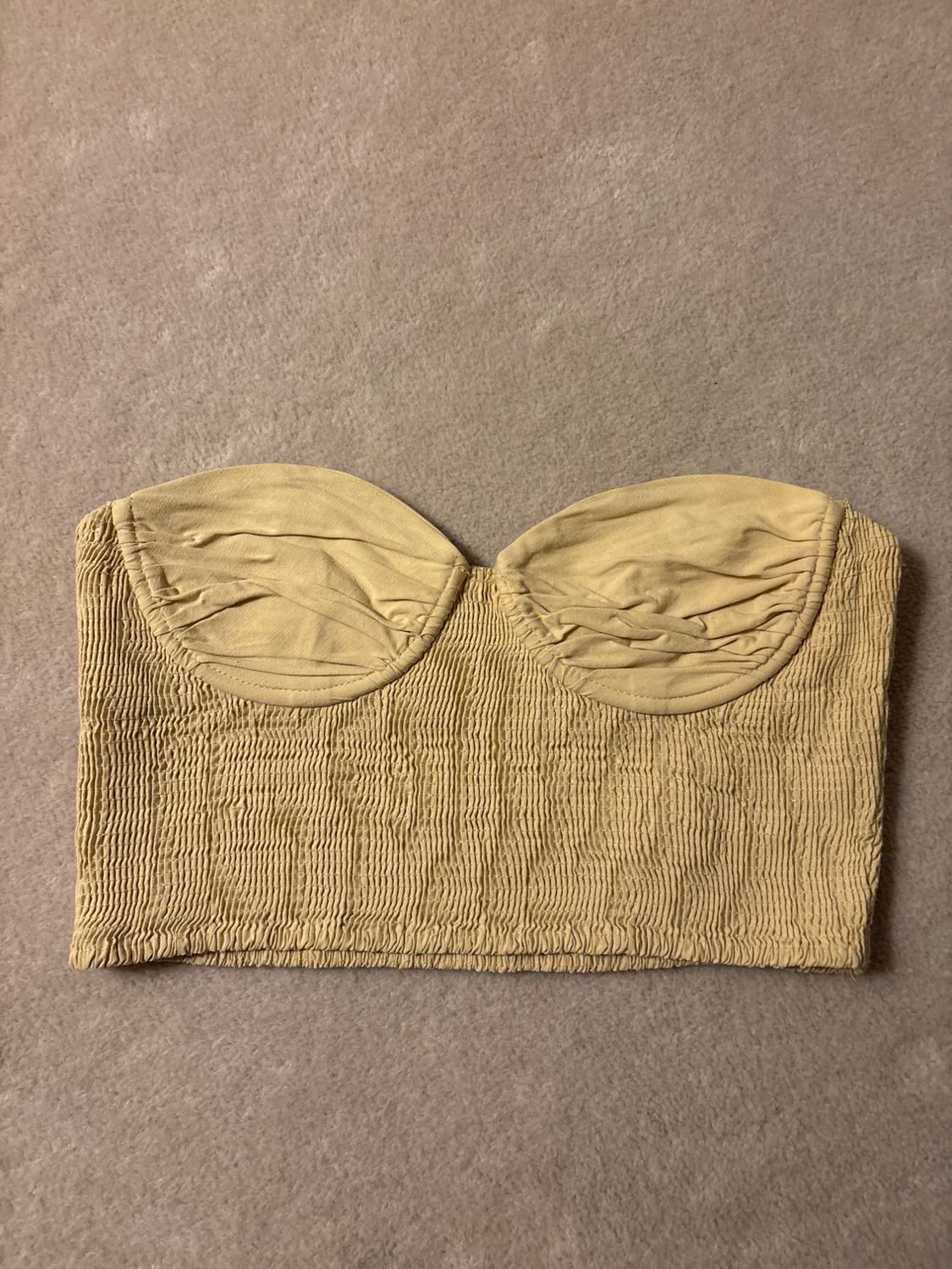 Top bandeau beige with ruched texture and draped cups, on a light grey fabric background.
