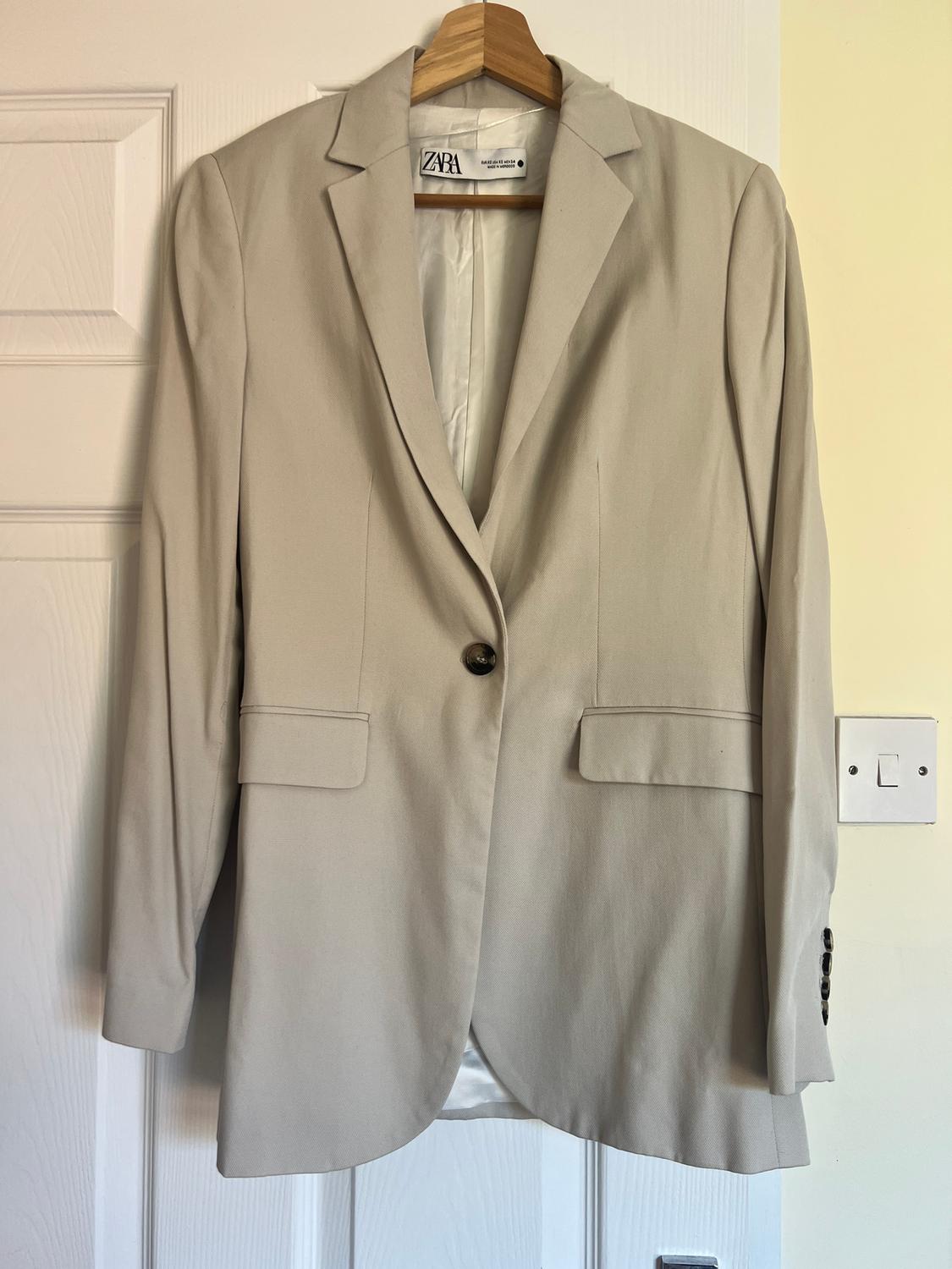 Beige jacket hanging on a wooden hanger, with a dark button and front pockets. Visible label: "ZARA". White door in the background.