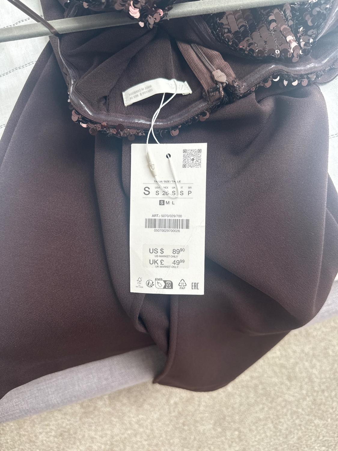 Brown clothing tag with sequins, showing sizes S, M, L and prices: US $89.90, UK £49.99. Item code visible.