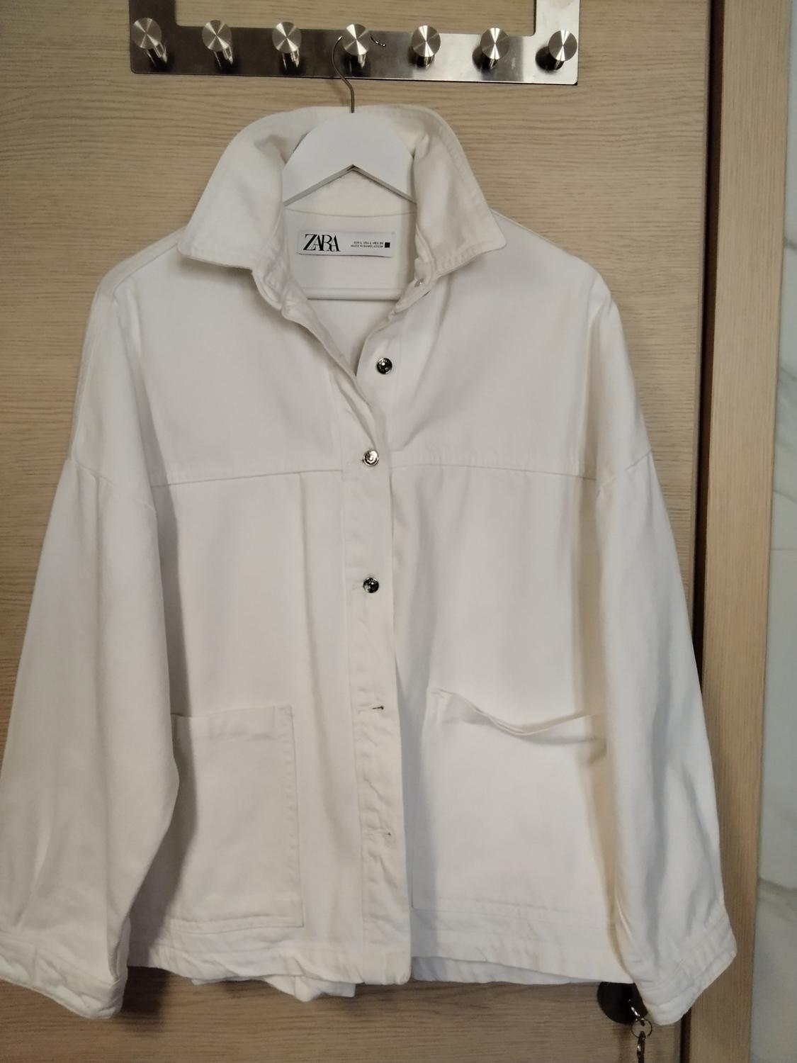 White jacket with press-stud buttons and pockets, hanging on a wooden door. Label with the text ZARA.